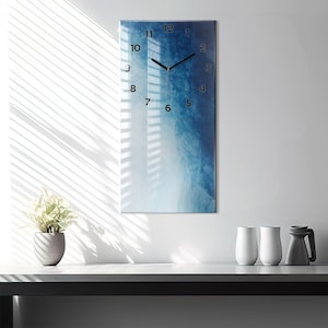 May include: A rectangular wall clock with a blue and white abstract design. The clock face features black numerals and hands. The clock is mounted on a white wall, with a plant in a gray pot and white vases on a table below.