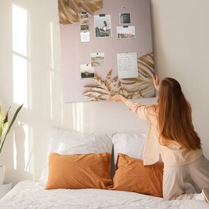 May include: A rectangular wall-mounted pin board featuring a light beige background with printed illustrations of dried palm leaves and pampas grass. The board displays several small photographs and a white note pinned to its surface with black clips.