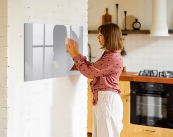 Puede incluir: Una persona está colgando una pizarra magnética rectangular gris en una pared de ladrillo blanco. La pizarra tiene un reflejo de ventana y una pequeña nota blanca pegada a ella.