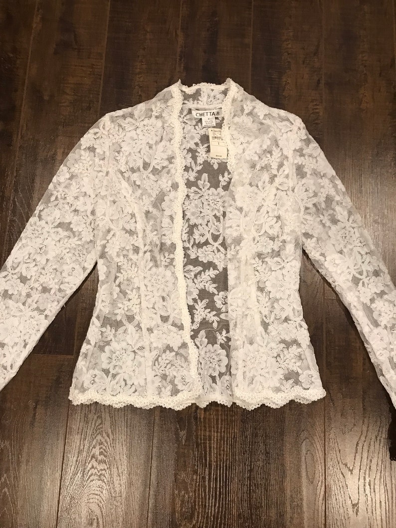 May include: A white lace jacket with long sleeves and a scalloped hem. The jacket is open in the front and features a floral pattern. The brand name "CHETTA B" is visible on a tag.