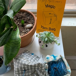 May include: A close-up of a white table with two gray and white yarn bags, one with a blue yarn ball inside. A small white planter with a succulent plant is in the background. A yellow sign with the text "Mary Mattimoe's Magic Yarn Bag" is leaning against a window.