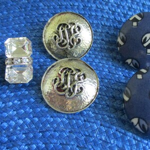 May include: A set of six earrings, including two pairs of silver tone earrings with a monogram design, and two pairs of blue fabric earrings with a white geometric pattern.