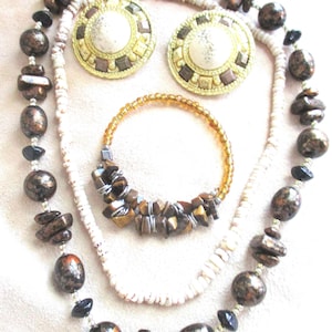 May include: A set of three necklaces featuring brown, white, and gold beads. The necklaces are layered and include a brown and gold beaded necklace, a white beaded necklace, and a brown beaded necklace with gold accents. The set also includes a pair of large, round, gold earrings with a white center and brown accents.