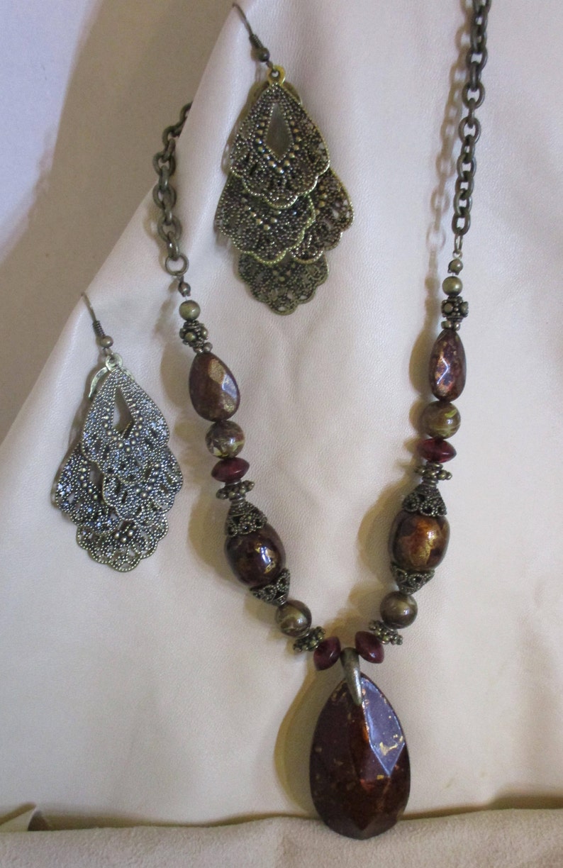 May include: A brown and gold necklace with a large teardrop-shaped pendant. The necklace is made of brown and gold beads and has a chain with a filigree design. The pendant is a teardrop shape with a brown and gold design. The necklace is on a white background.
