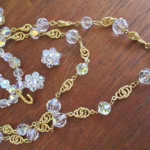 May include: A gold chain necklace with clear crystal beads. The necklace is layered with a second strand of smaller, iridescent beads. There are also two small clusters of clear crystal beads.