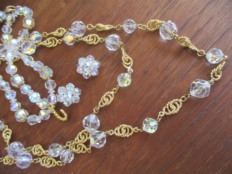 May include: A gold chain necklace with clear crystal beads. The necklace is layered with a second strand of smaller, iridescent beads. There are also two small clusters of clear crystal beads.
