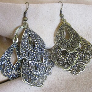 May include: A pair of gold-toned dangle earrings with a layered, intricate design. Each earring features multiple tiers of delicate, scalloped shapes with a textured finish.