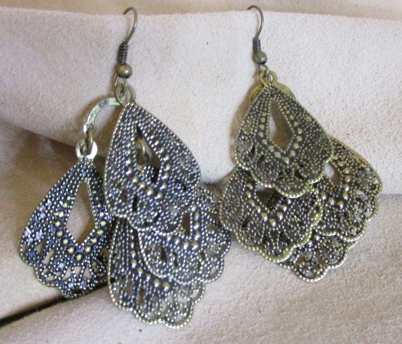 May include: A pair of gold-toned dangle earrings with a layered, intricate design. Each earring features multiple tiers of delicate, scalloped shapes with a textured finish.