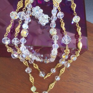 May include: A three-strand necklace with gold chain and clear crystal beads. The necklace is displayed on a purple velvet box.