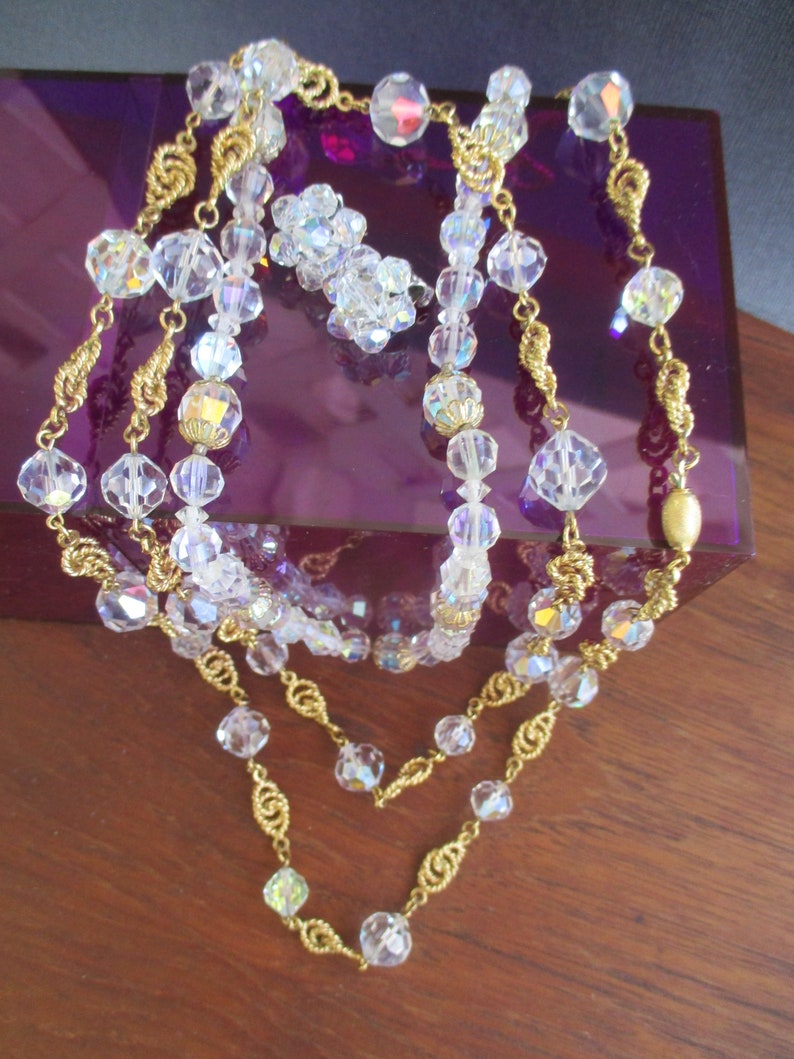 May include: A three-strand necklace with gold chain and clear crystal beads. The necklace is displayed on a purple velvet box.