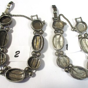 May include: Two silver tone bracelets with oval-shaped charms. The charms have a textured design and are connected by a chain. The bracelets have a clasp closure. The bracelets are labeled "1" and "2".