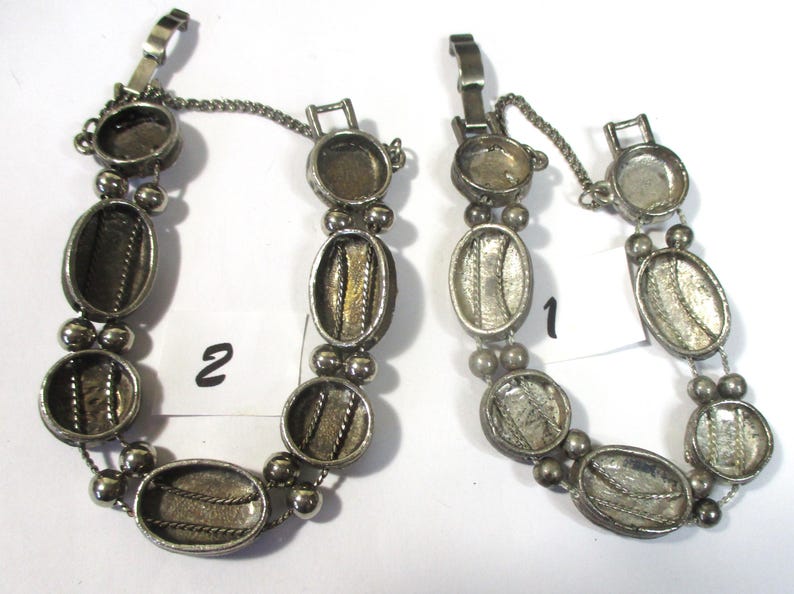 May include: Two silver tone bracelets with oval-shaped charms. The charms have a textured design and are connected by a chain. The bracelets have a clasp closure. The bracelets are labeled "1" and "2".