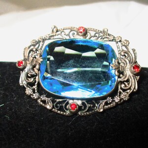May include: A silver filigree brooch with a large, faceted, blue glass stone in the center. The brooch is accented with four small, red glass stones.