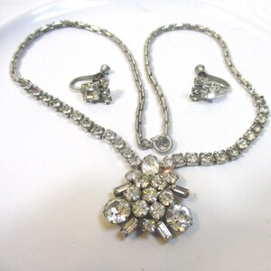 May include: A vintage silver-toned necklace and matching clip-on earrings set. The necklace features a central cluster of clear, sparkling rhinestones and a delicate chain. The earrings have a similar design. The jewelry set is displayed on a white surface.
