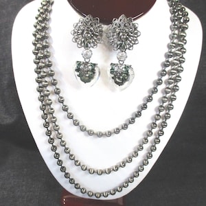 May include: A three-strand silver necklace with round beads and a pair of silver earrings with a floral design and a clear heart pendant.