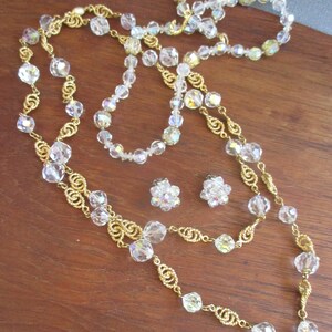 May include: A set of iridescent crystal necklaces and earrings with gold chain accents. The necklaces are layered and feature a variety of crystal shapes. The earrings are small and feature a cluster of crystals.