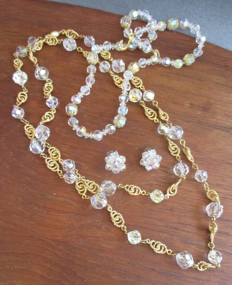 May include: A set of iridescent crystal necklaces and earrings with gold chain accents. The necklaces are layered and feature a variety of crystal shapes. The earrings are small and feature a cluster of crystals.