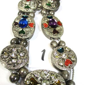 May include: A silver bracelet with multiple oval-shaped charms. Each charm is decorated with colorful enamel and rhinestones. The charms are connected by a silver chain and silver beads.