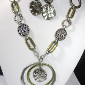 May include: A silver and gold necklace with a large circular pendant and matching earrings. The pendant is made of two overlapping circles, one silver and one gold, with a textured silver disc in the center. The earrings are simple silver discs with a textured surface.