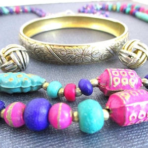 May include: A set of jewelry featuring a silver bangle with an engraved floral design, a pair of silver knot earrings, and a multi-colored beaded necklace with pink, turquoise, and purple beads.