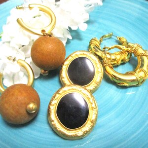 May include: Three pairs of gold tone earrings. One pair features a large brown wooden bead, another pair has a black enamel circle, and the third pair is a thick gold hoop with a braided design.