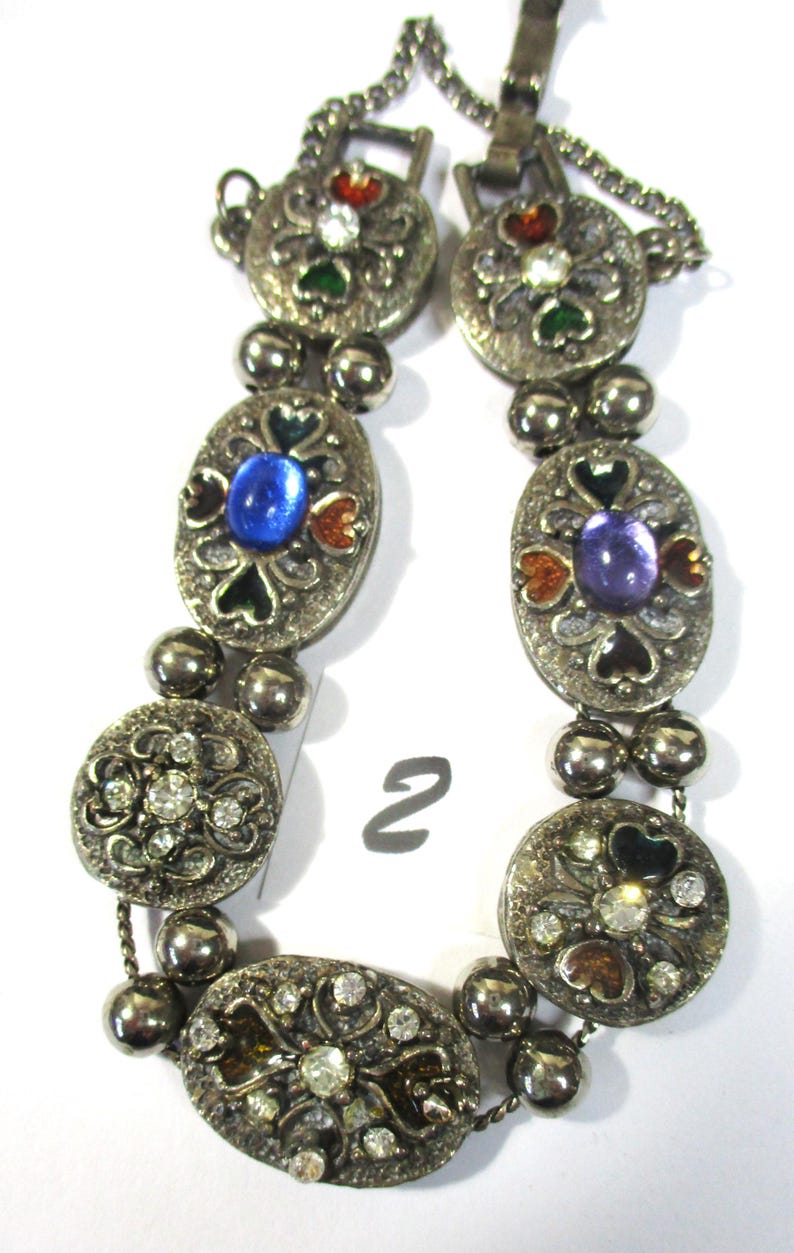 May include: A silver bracelet with oval-shaped charms featuring colorful enamel and rhinestones. The charms are connected by silver beads and a silver chain.