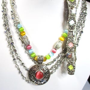 May include: A multi-strand necklace with a silver pendant featuring a pink stone. The necklace is made of silver chains and colorful beads.