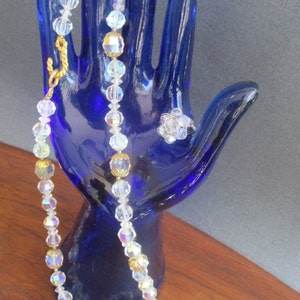 May include: A crystal necklace with gold accents and two matching crystal rings. The necklace and rings are displayed on a blue hand-shaped mannequin.