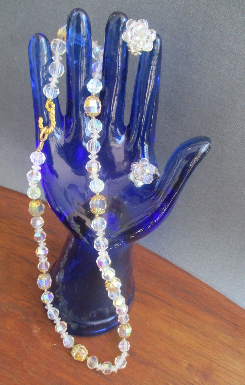 May include: A crystal necklace with gold accents and two matching crystal rings. The necklace and rings are displayed on a blue hand-shaped mannequin.
