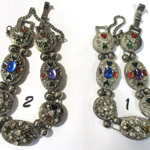 May include: Two silver bracelets with colorful enamel and rhinestone accents. The bracelets are linked together with a chain. The bracelets are numbered 1 and 2.