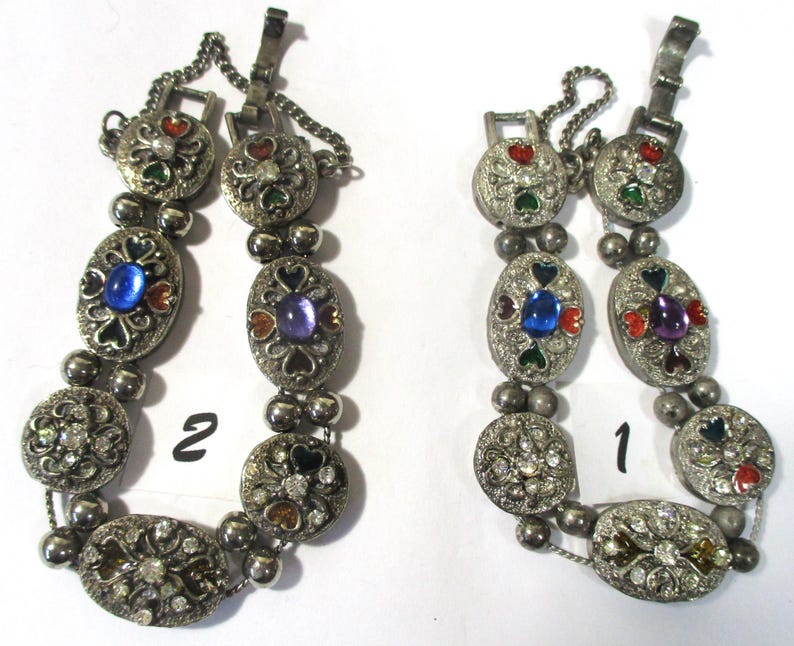 May include: Two silver bracelets with colorful enamel and rhinestone accents. The bracelets are linked together with a chain. The bracelets are numbered 1 and 2.