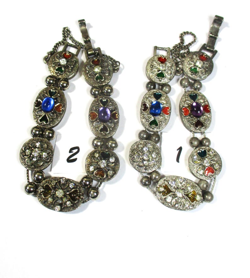 May include: Two silver bracelets with colorful enamel and rhinestone accents. The bracelets have a heart design and are numbered 1 and 2.