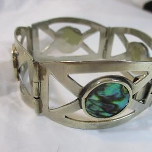 May include: Silver-toned hinged bracelet with geometric cutouts and oval abalone shell inlays. The bracelet has a clasp closure and is designed with a series of connected panels. The abalone shell displays iridescent green and blue hues.