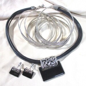 May include: A black leather necklace with a silver pendant and matching earrings. The pendant is a rectangular shape with an intricate silver design and a black stone in the center. The earrings are small and have a similar design to the pendant.