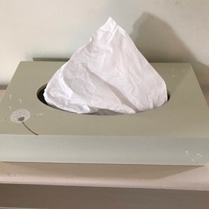May include: A light green tissue box cover with a white tissue inside. The cover features a white dandelion design with seeds blowing in the wind.