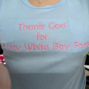 May include: Light blue t-shirt with the embroidered text "Thank God for Slutty White Boy Rock" in pink. The shirt has short sleeves and a crew neck.