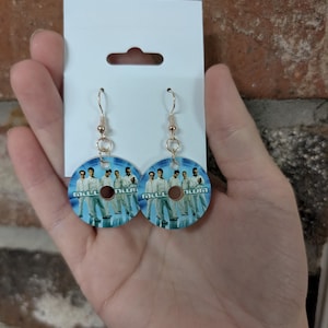 May include: A pair of circular dangle earrings featuring a graphic print of the Backstreet Boys from their Millennium album cover. The earrings have gold-toned metal hooks and are attached to a white rectangular display card with a cutout hole.