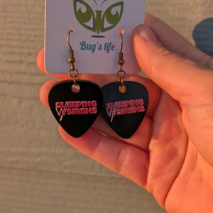 May include: A pair of black guitar pick earrings featuring the text Sleeping With Sirens in pink lettering. The earrings hang from bronze-toned metal hooks and are attached to a white display card with a green logo and the text Bug's life.