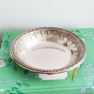 May include: A silver-colored, round bowl with an ornate, detailed rim. The bowl has a reflective surface and sits on a green surface with floral patterns. The rim features a repeating pattern of decorative elements.