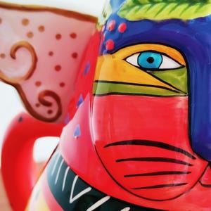 May include: Close-up of a vibrantly painted ceramic pitcher. The pitcher features a stylized face with a blue eye, red mouth, and colorful accents. The ear is pink with brown swirls and dots. The design is bold and artistic.