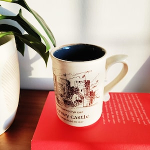 May include: Cream-colored ceramic mug with a dark blue interior and a curved handle. The mug features a sepia-toned illustration of a castle with the text "Conwy Castle completed in 1287". The mug is resting on a red book.