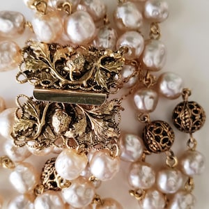 Puede incluir: Primer plano de un collar vintage con múltiples hilos de perlas blanquecinas de forma irregular. El collar tiene un cierre dorado con intrincados detalles de hojas y bayas. Las perlas son de color crema y el cierre añade un toque de elegancia.