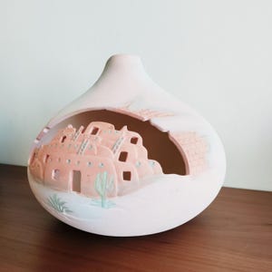 Puede incluir: Un jarrón de cerámica rosa claro con un diseño del suroeste. El jarrón presenta una sección recortada que muestra un relieve detallado de edificios de adobe, un cactus y otros elementos del desierto. El jarrón es un objeto decorativo.