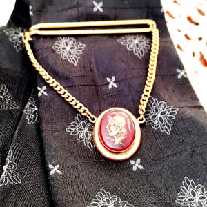 May include: Gold-toned tie clip with a chain and a red oval pendant. The pendant features a white profile of a person in a helmet. The clip is on a black tie with a white floral pattern.