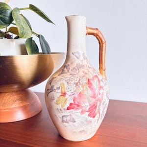 May include: A cream-colored ceramic pitcher featuring a floral pattern with red, yellow, and grey leaves and blossoms. The vessel has a narrow neck and a metallic copper-toned handle shaped like a bamboo stalk, extending from the rim to the shoulder.