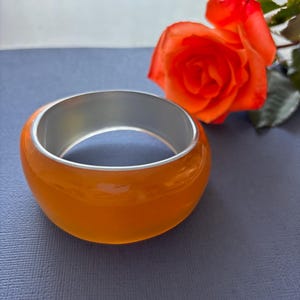 May include: Orange bangle bracelet with a silver interior. The bracelet is smooth and glossy. A vibrant orange rose is in the background, set against a blue surface.