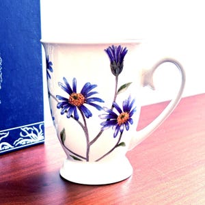 May include: A white ceramic mug featuring a botanical illustration of blue daisy-like flowers with yellow centers and green stems. The mug has a flared rim, a pedestal base, and a curved handle. The design is painted on the exterior surface.