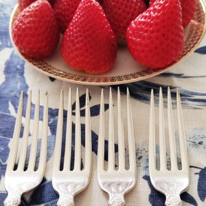 Könnte beinhalten: Ein Teller mit reifen roten Erdbeeren mit dekorativem Rand liegt über vier Silberbesteckgabeln. Die Gabeln sind auf einer blau-weiß geblümten Tischdecke angeordnet. Die Erdbeeren sind frisch und lebendig.