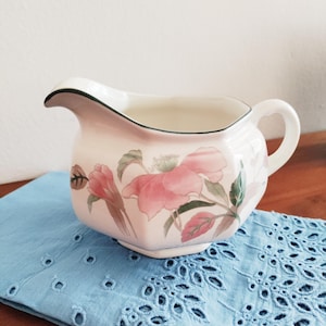 May include: A vintage cream pitcher with a floral design. The pitcher is off-white with pink flowers and green leaves. It has a black trim and a handle. The pitcher is sitting on a blue eyelet cloth.