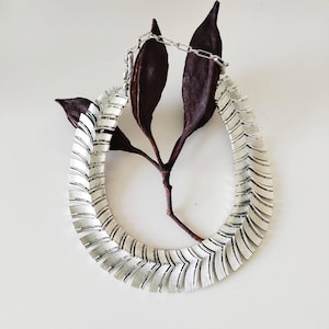 May include: A silver-toned collar necklace featuring a series of overlapping, chevron-shaped metal plates with a brushed finish. The necklace is constructed with a flexible chain link design and includes a thin metal chain extension at the top.